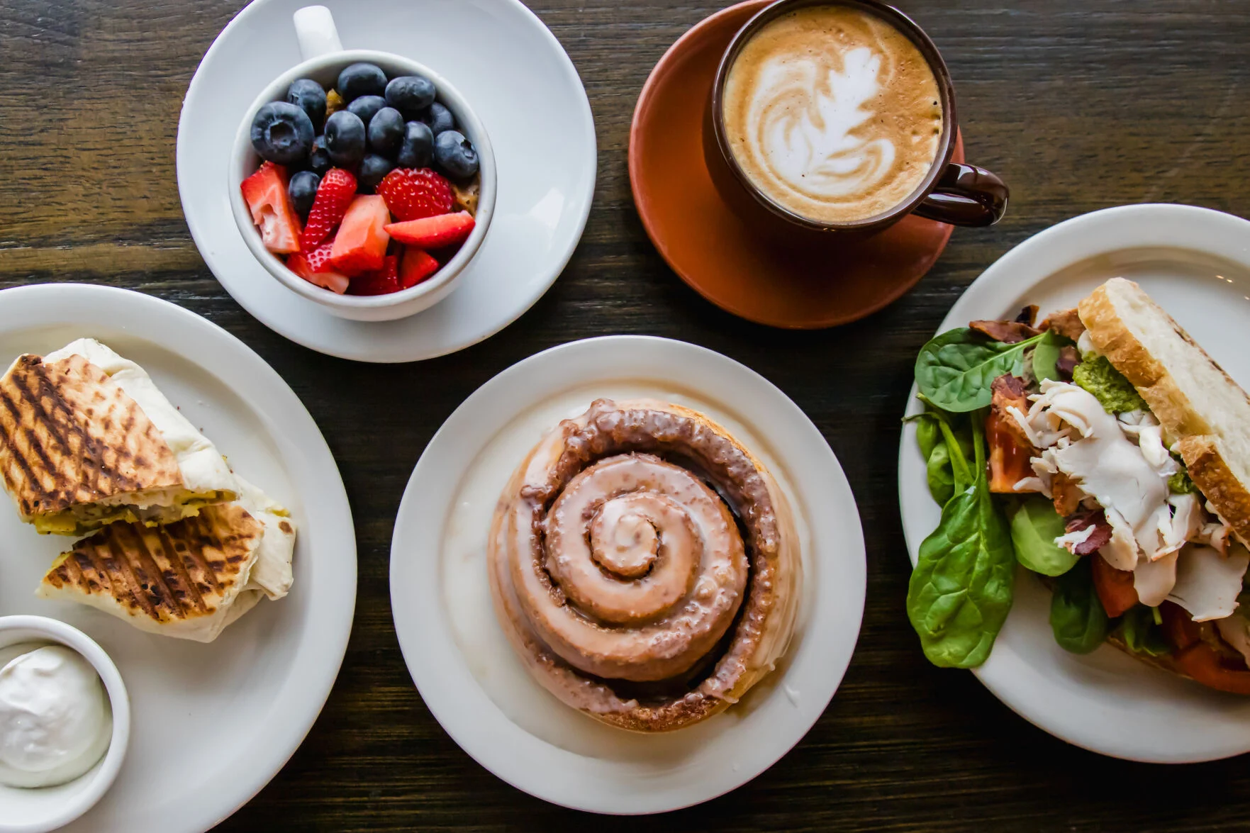 A breakfast spread at 222: breakfast burrito, fruit cup, latte, cinnamon roll, and a BLT